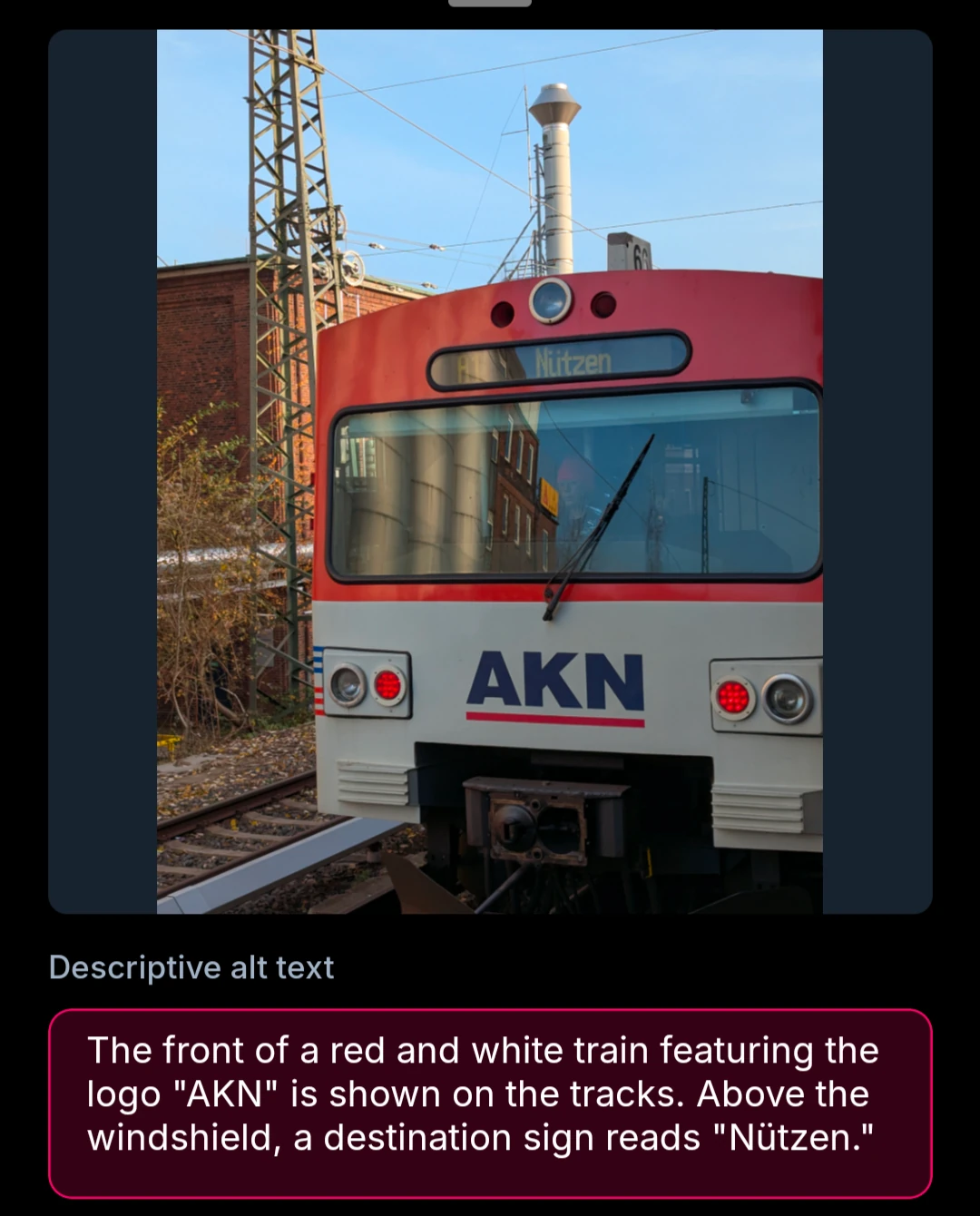The front of a red and white train featuring the logo "AKN" is shown on the tracks. Above the windshield, a destination sign reads "Nützen."