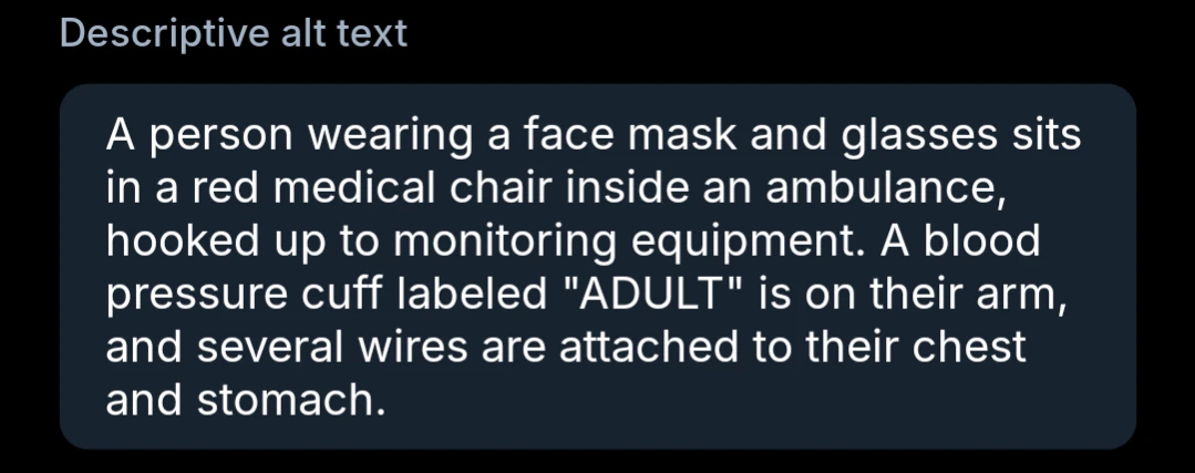A person wearing a face mask and glasses sits in a red medical chair inside an ambulance, hooked up to monitoring equipment. A blood pressure cuff labeled "ADULT" is on their arm, and several wires are attached to their chest and stomach.