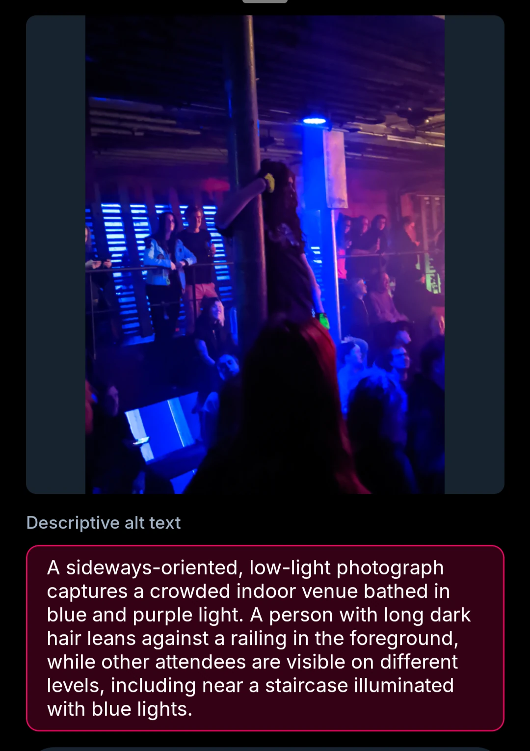 A sideways-oriented, low-light photograph captures a crowded indoor venue bathed in blue and purple light. A person with long dark hair leans against a railing in the foreground, while other attendees are visible on different levels, including near a staircase illuminated with blue lights.
