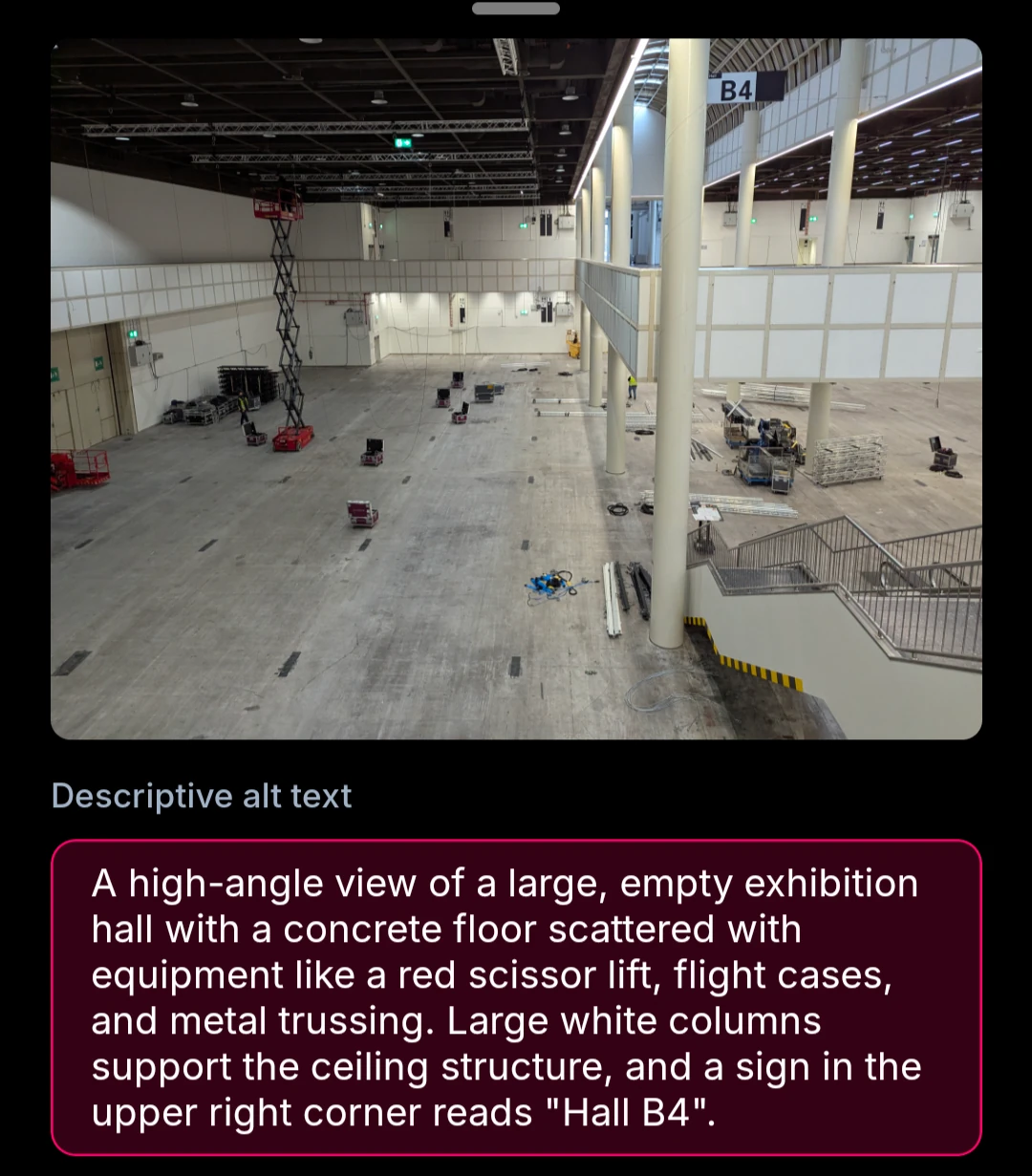 A high-angle view of a large, empty exhibition hall with a concrete floor scattered with equipment like a red scissor lift, flight cases, and metal trussing. Large white columns support the ceiling structure, and a sign in the upper right corner reads "Hall B4".