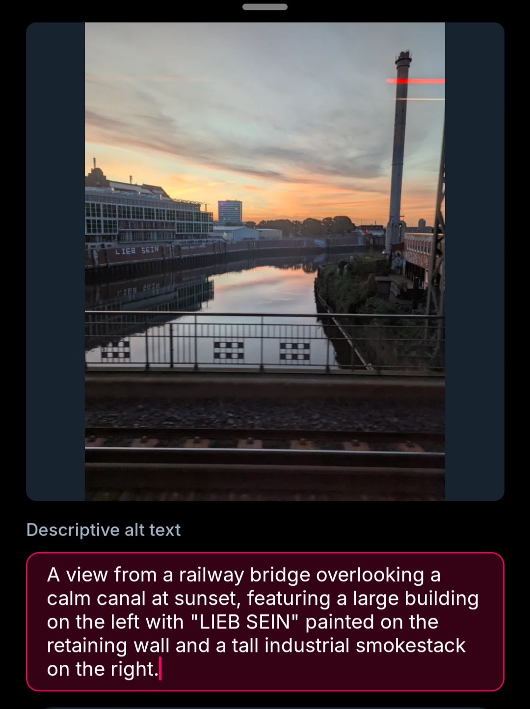 A view from a railway bridge overlooking a calm canal at sunset, featuring a large building on the left with "LIEB SEIN" painted on the retaining wall and a tall industrial smokestack on the right.