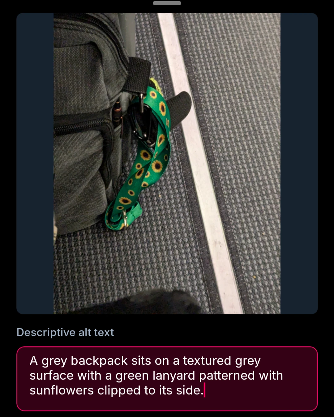 A grey backpack sits on a textured grey surface with a green lanyard patterned with sunflowers clipped to its side.