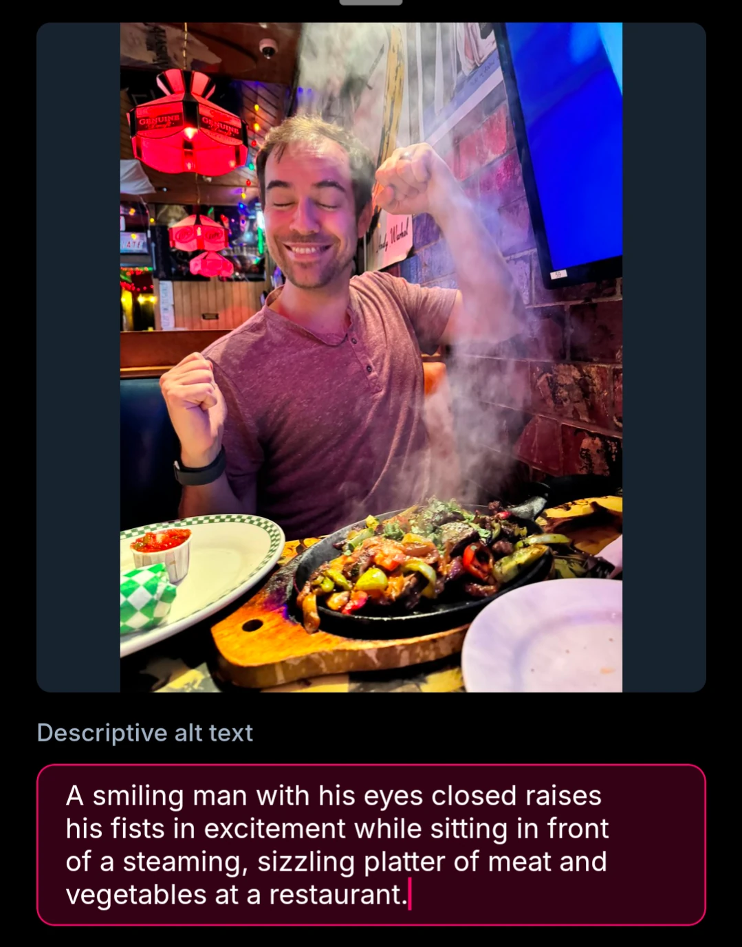 A smiling man with his eyes closed raises his fists in excitement while sitting in front of a steaming, sizzling platter of meat and vegetables at a restaurant.
